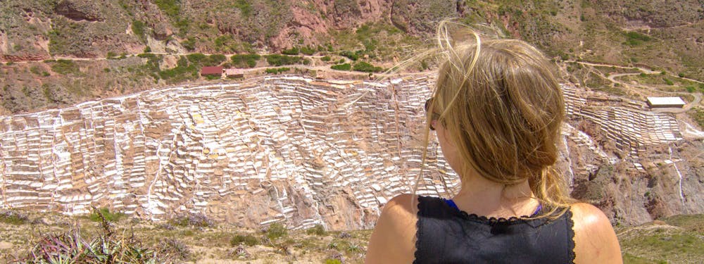 Best Hikes In Peru Image: A woman with a blonde braid pulled over her right shoulder is wearing a black top, sunglasses, and is observing the salt mines from across the valley.