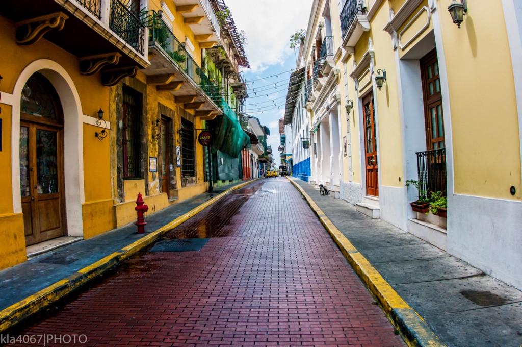 Architecture In Panama Image: A narrow city street is lined with buildings on both the left and right. We can see shades of mustard yellow, pale lemon, turquoise, and marine blue gracing their faces. Many have wrought iron balconies.