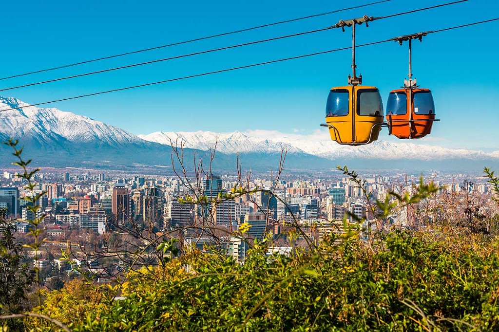 Destinations Worth Dreaming Image: An enclosed aerial tram has two pods—one yellow, one orange. They are sailing above the city of Chile; a snow-capped mountain range sits in the background, the city skyline is below, and greenery graces the foreground.