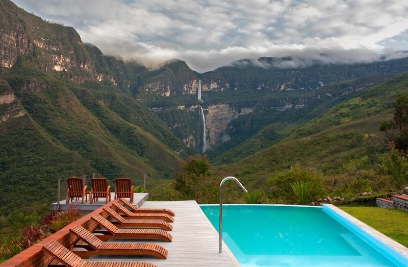 Best Hikes In Peru Image: A hotel swimming pool is surrounded by wooden lounge chairs, and offers an incredible view of the waterfall at the other end of this mountainous landscape.