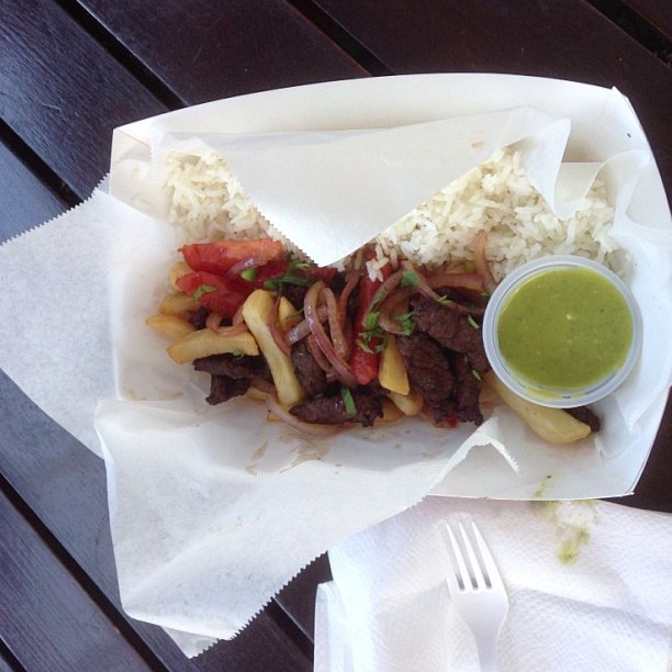 Visit Peru Image: A to-go basket is filled with the colourful beef and vegetable stir-fry has fluffy white rice on the side, and fried potatoes throughout. A green dipping sauce sits in the basket, and a napkin and white plastic fork are off to the side.