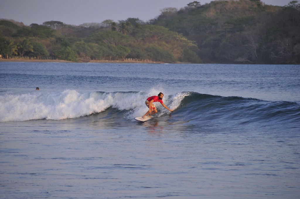 Best Surfing Beaches: A surfer wearing a red wetsuit top and red and white swim shorts rides a surf board, and touches his hand to a cresting wave.