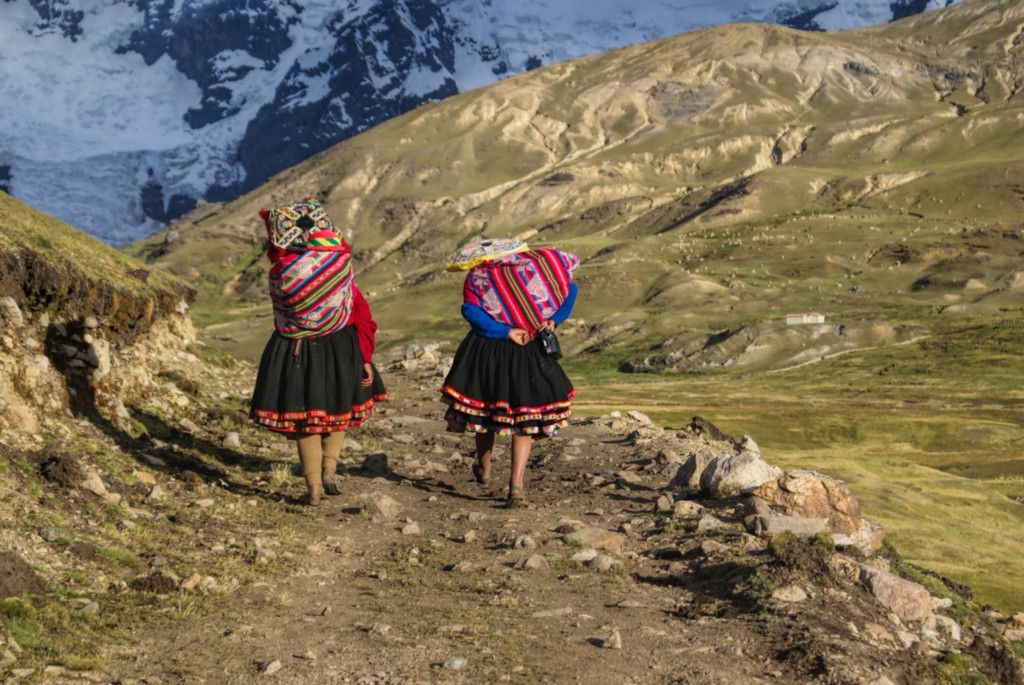 Central and South American Ruins Image: Two women have their backs to us. They are wearing colourful skirts, and are carrying equally colourful bundles on their backs as they walk down an incline in the Andes. 