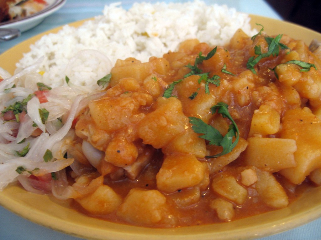 Visit Peru Image: The hearty potato stew described in this section of our Peru food tour is heaped onto a yellow plate, and accompanied by white rice, and what appears to be a vegetable slaw.