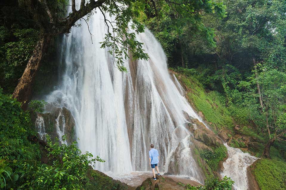 Destinations Worth Dreaming Image: A male traveller wearing dark-blue shorts and a light-blue shirt stands on a rock observing a waterfall as it rushes and breaks down a slope in—streaming into white channels.