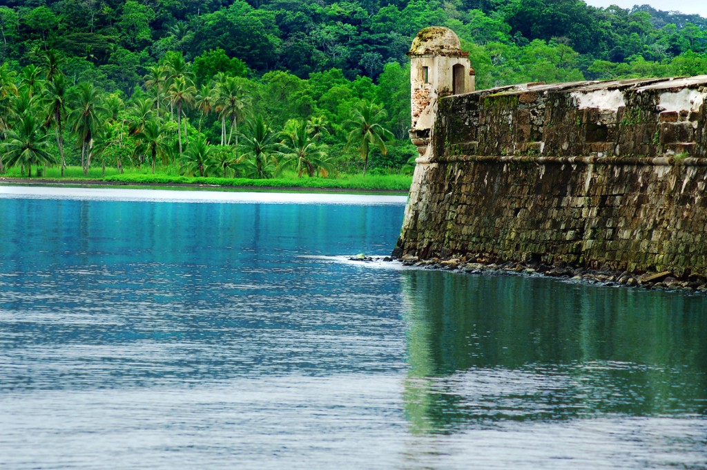 Architecture In Panama Image: A fort aged and in the early stages of being reclaimed by natures has openings which overlook calm blue waters, and a vivd green forest.