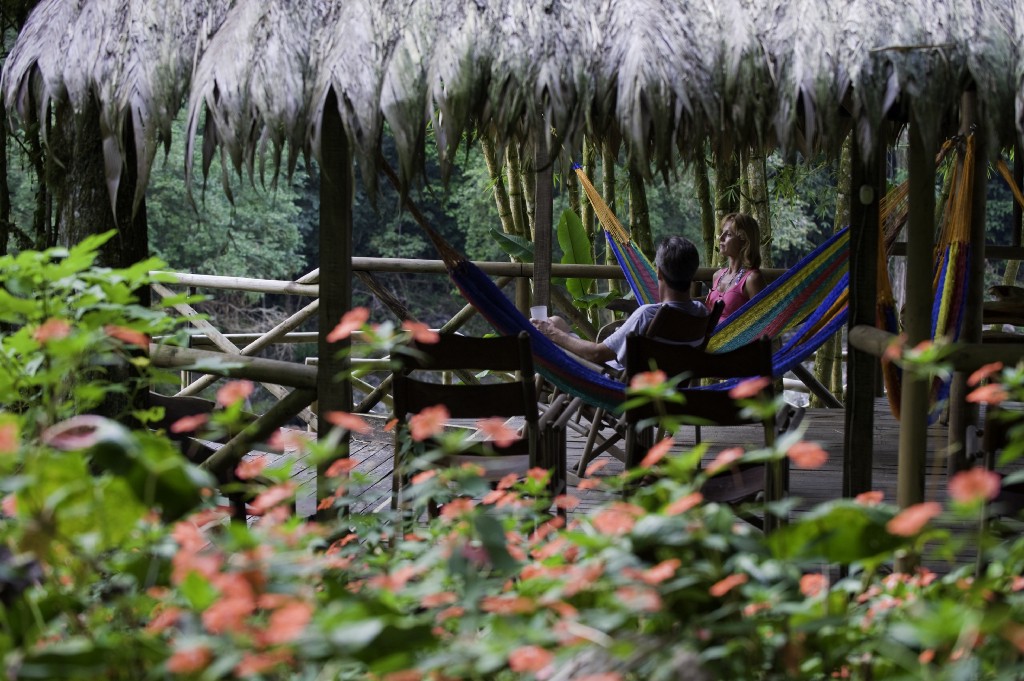 Eco-Friendly Costa Rica Image: A couple sits in two hammocks in an open-air structure covered by a leafy roof. They are talking and enjoying the surrounding jungle scenery.