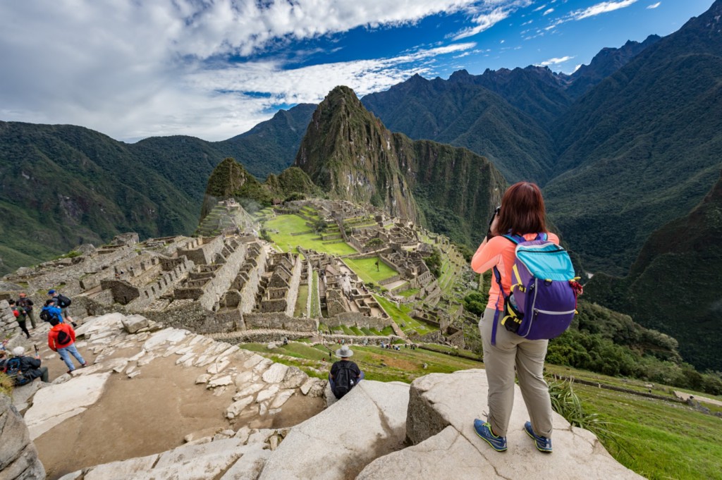 Central and South American Ruins Image: Travellers are staggered on different levels of a ruin at Machu Picchu. At the top most level, a woman wearing a backpack is poised to take a photograph.