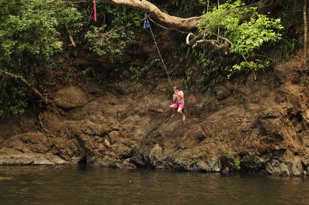 Swimming Holes Image: A young man in red swim shorts prepares to jettison himself off of rocks to swing on a rope suspended from a tree branch, and plunge into the water below.