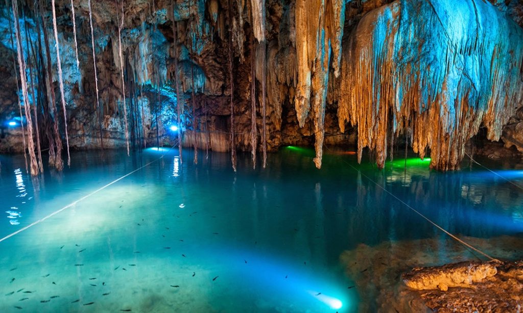 Cave Floating Image: Stalactites drop down from the top of a cavern—hovering above turquoise water which is illuminated from below. Fish can be seen swimming.