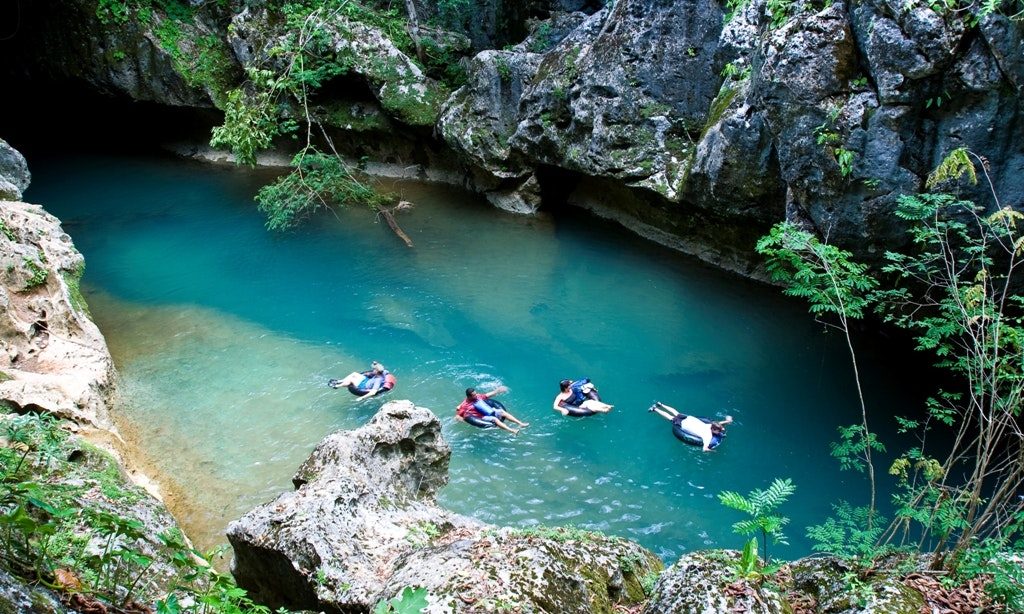 Moving to Belize Image: An aerial view of four tubers emerging from a cave and floating into a beautifully blue pool surrounded by jungle.