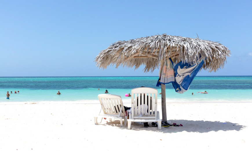 Are Cuba's Beaches Nice Image: A leafy beach umbrella strewn with a towel shades two chairs on the beach.