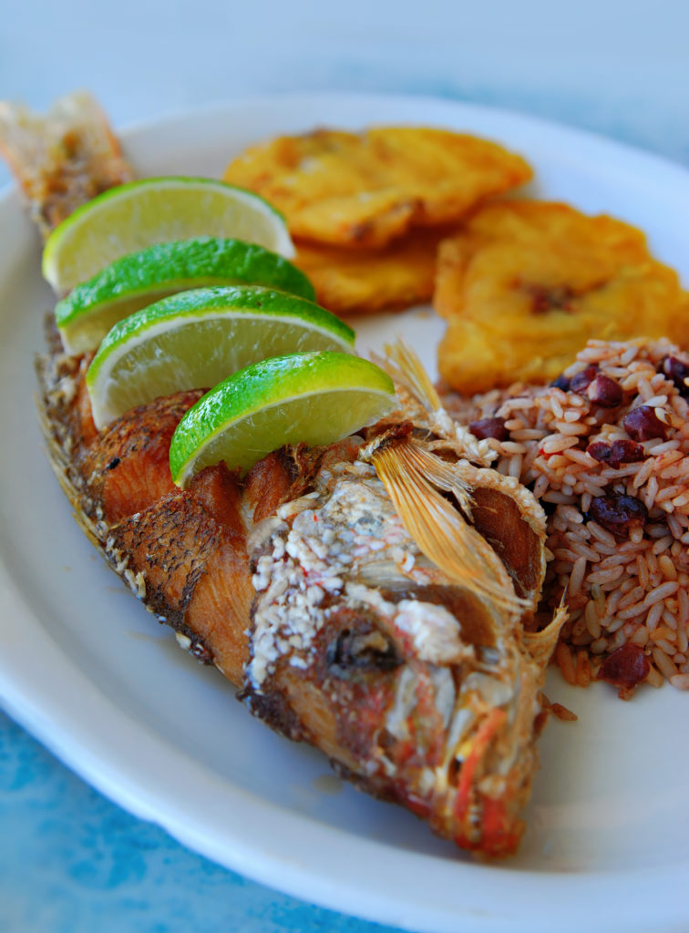 Costa Rican Foods Image: A serving of fried red snapper with pinto beans, rice, and tostones (Cuban plantains). Four lime wedges have been placed inside of slits made in the side of the fish.