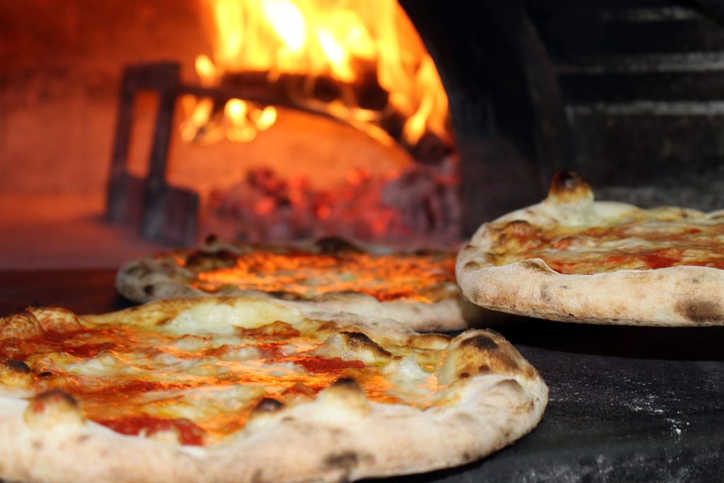 Costa Rican Foods Image: Three pizzas sit in the foreground whilst the open mouth of the pizza oven glows with fire in the background.