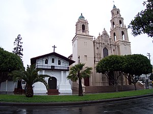 Cuba Unusual Museum Image: A photograph shows a smaller white chapel with a black roof, and the larger main cathedral on a tree-lined street.