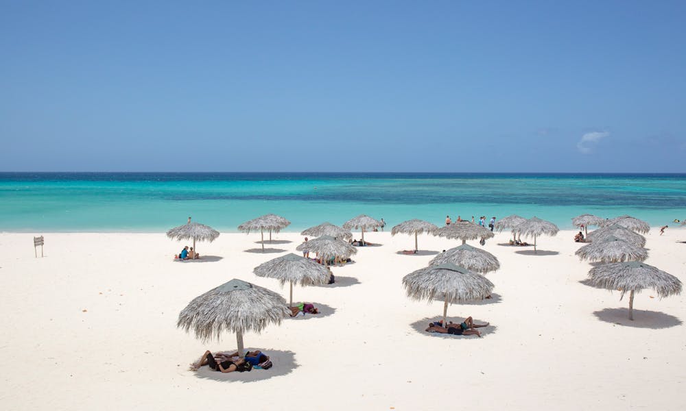 Best Beaches Image: Cuba. A series of straw beach umbrellas creates circles of shade in the sand for beachgoers to cool off in. The sand is pristine, the water is inviting, and there is hardly a cloud in the sky.
