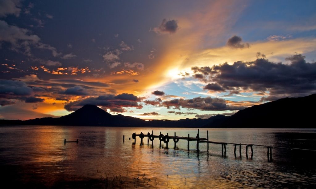 Mayan Image: A pier leading up to Guatemala's Lake Atitlán is caressed by a sun dappled sky in shades of blue, gold, and purple.