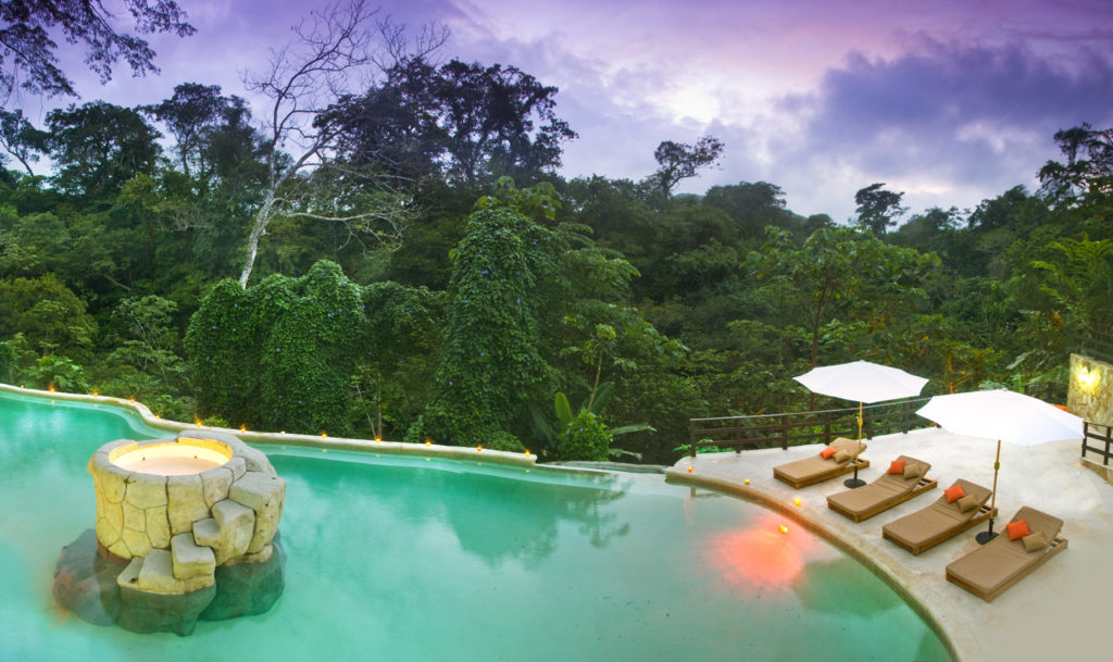 Hot Springs Image: Hot Springs, Tizate. Beige lounge chairs and white umbrellas line a beautiful pool, which is in fact a hot spring. The background is a jungle and a purple sky.