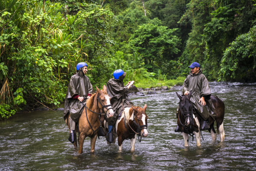 Costa Rica Adventure Trips Image: A woman takes a selfie on a white and brown horse while crossing the river Arenal on horseback near the small town of La Fortuna, in Costa Rica.