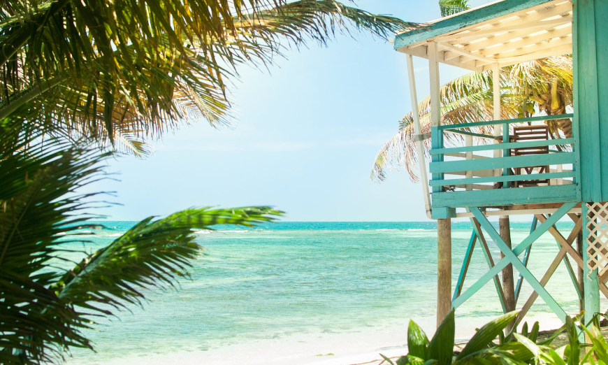 Belize Island Image: Side view of a colorful stilted bungalow, palm fronds, and tranquil shoreline. 