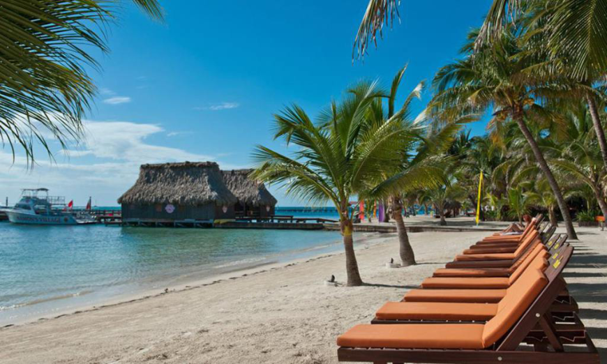 Best Beaches for Swimming in Belize Image: A strip of Ramon's beach is dotted with a few palm trees. Wooden lounge chairs are in the foreground. Thatched roof bungalows are in the background.