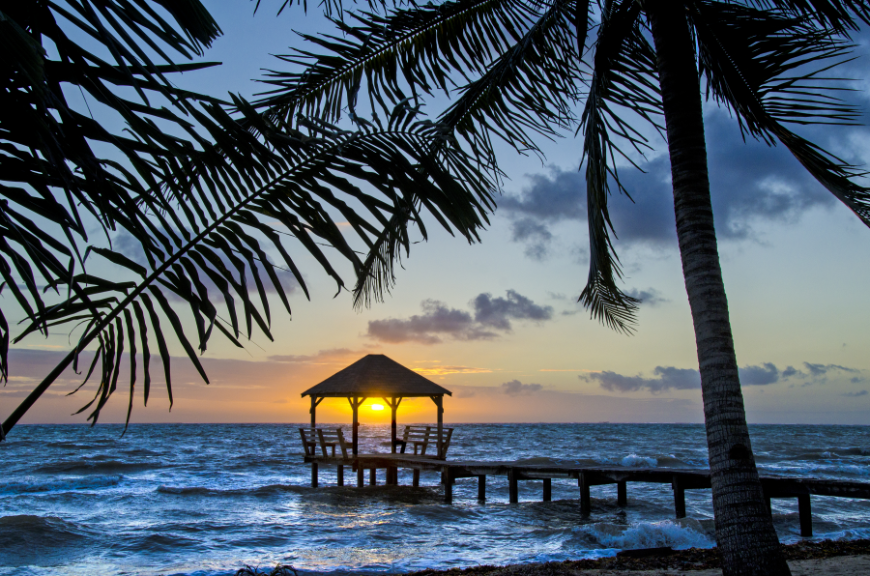 Best Islands in Belize for Everyone Image: An open-air thatched hut with chairs sits at the bottom of a pier at sunset.