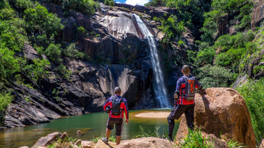 Best time to visit Central America Image: Two men with shaved heads have their backs to the camera as they look on at a waterfall; they are dressed in long sleeved outdoor apparel, and are carrying backpacks.