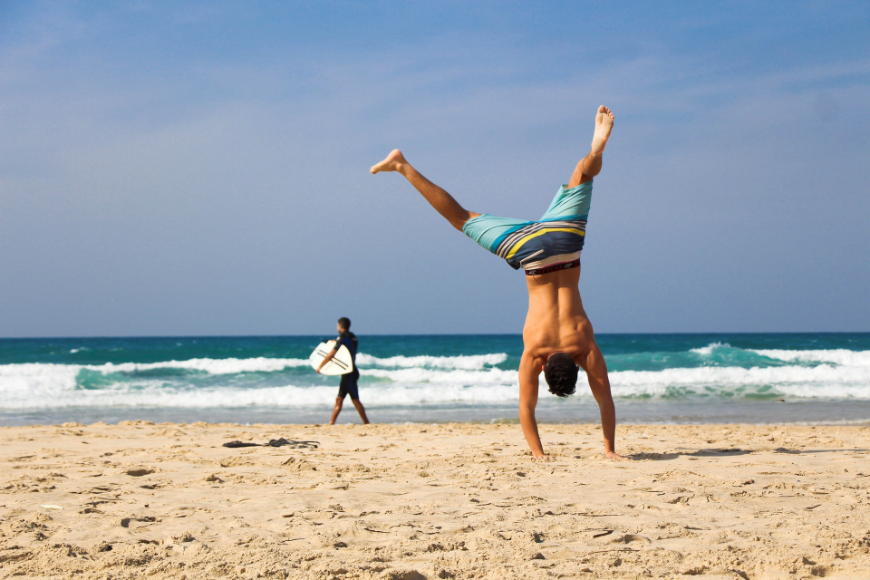 Moving to Belize Image: A man in striped swim shorts does a cartwheel in the sand, while a surfer in a wetsuit approaches the water in the background.