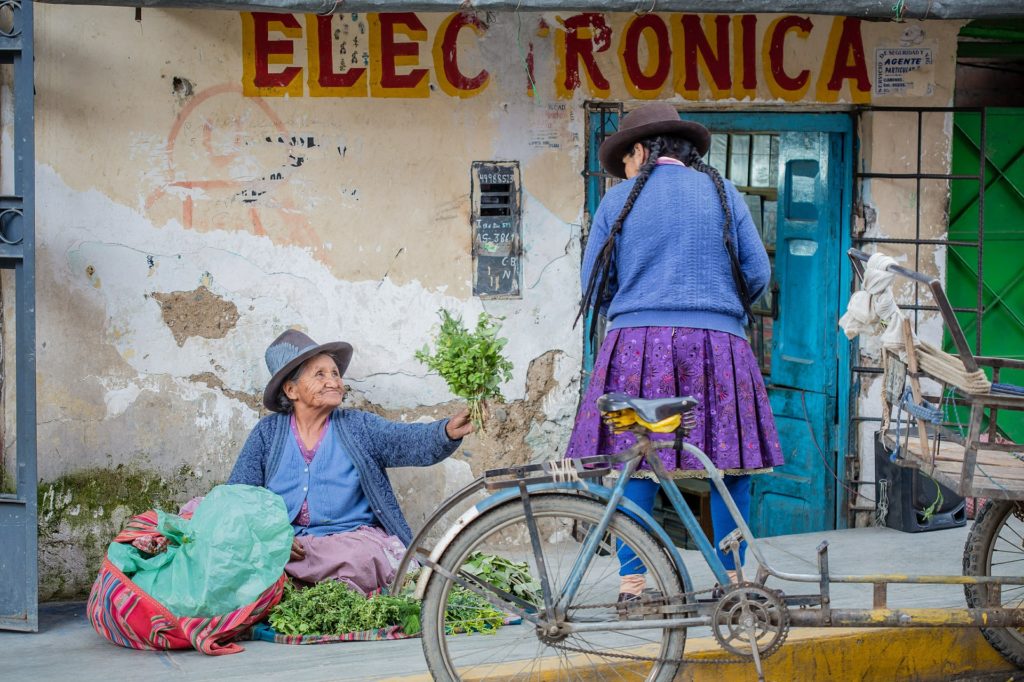 Best Time To Visit South America Image: An elder Peruvian woman sits on a sidewalk and proffers a bundle of fresh greenery to a younger woman.