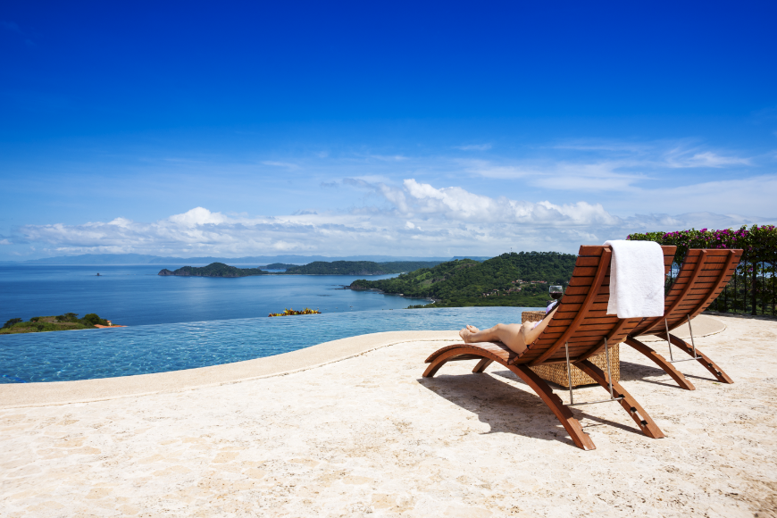 Best Travel Stories Image: Two chairs overlook an immaculately blue sky and water; bits of greenery are seen from nearby islands, and gentle white clouds hang low in the sky.