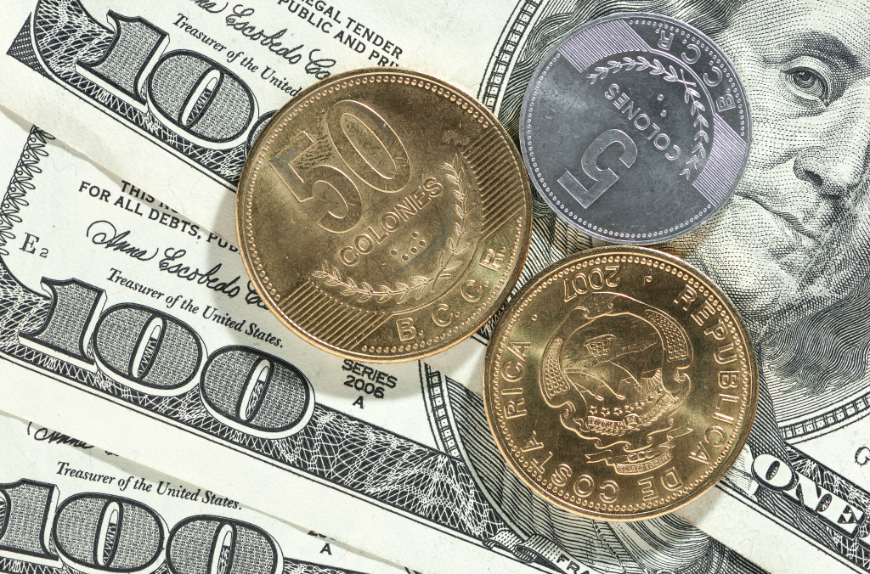 Booking A Vacation Online Image: Three Costa Rican Colónes coins (two gold, one silver) sit atop three one hundred United States Dollar bills. 