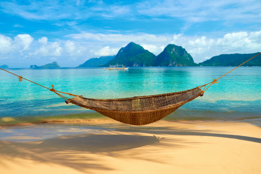 Booking A Vacation Online Image: A hammock is suspended on a tropical beach over golden sand. Clear turquoise waters, a blue and white sky, and green mountainous islands are seen in the distance.