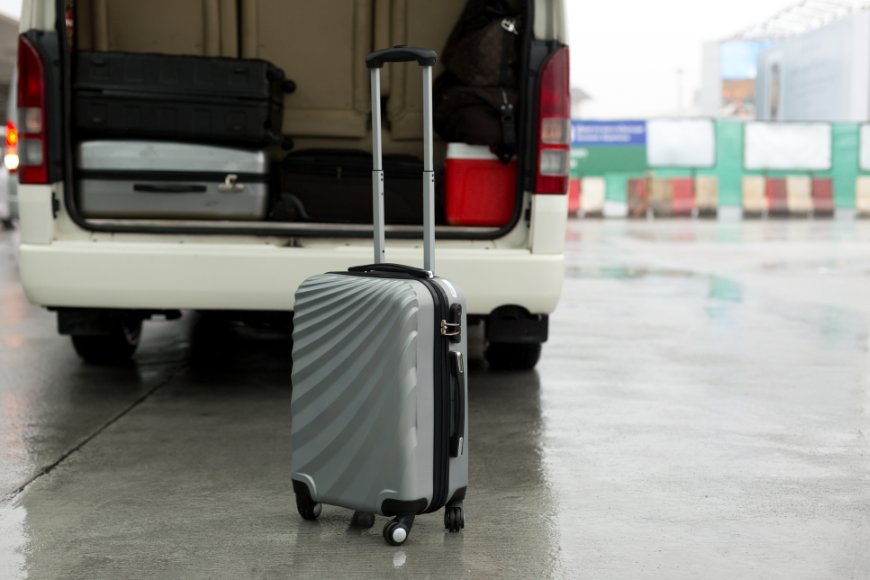 Booking A Vacation Online Image: A silver suitcase sits outside of the trunk of a white van already loaded with other suitcases.