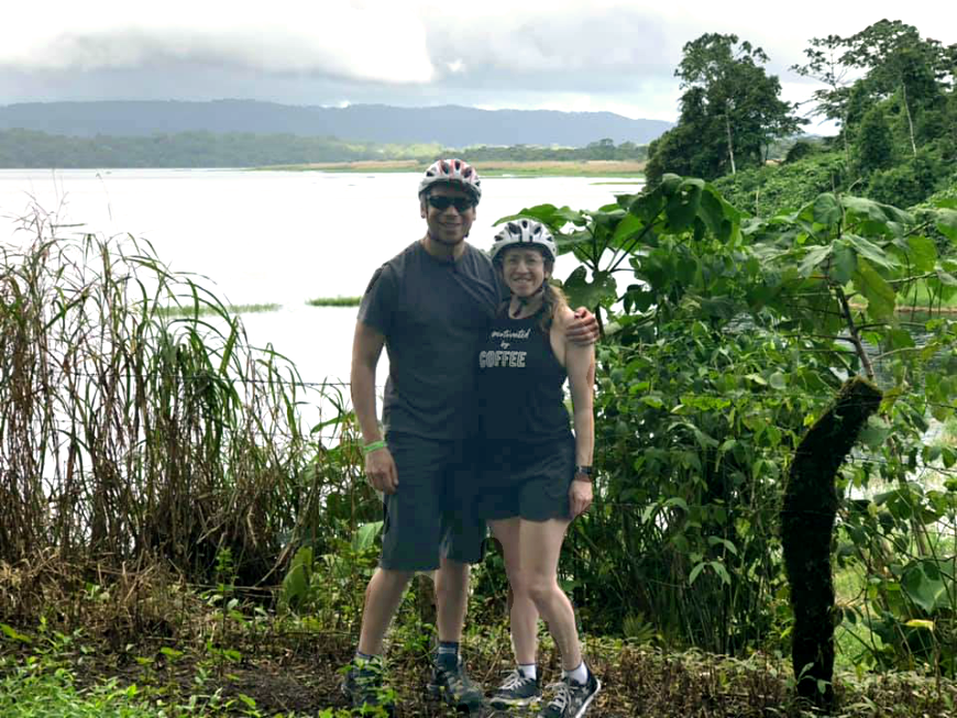 Costa Rica Couples Trip: The Pearson/Lepine's smile in front of Lake Arenal wearing bike helmets.