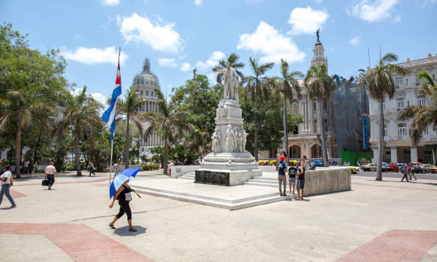 Cuban Homestays Image: A woman walks through a plaza under an umbrella. There is a statue and a Cuban flag.