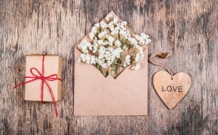 Eco-Friendly Destination Wedding Image: Brown box with a red string bow. A wooden heart with love written on it. A brown paper envelope full of white flowers.