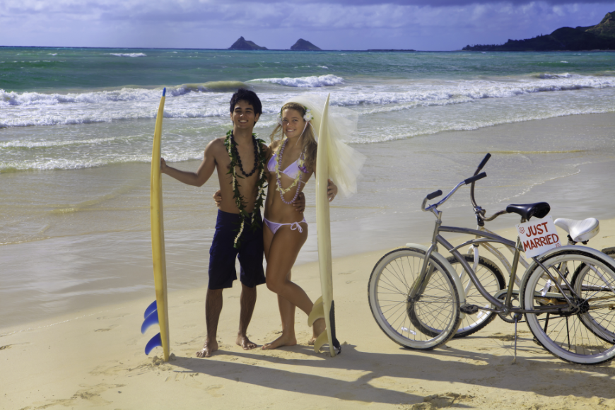 Eco-Friendly Destination Wedding Image: Newlyweds pose in swimsuits holding yellow surfboards; bikes are parked off to the side. 