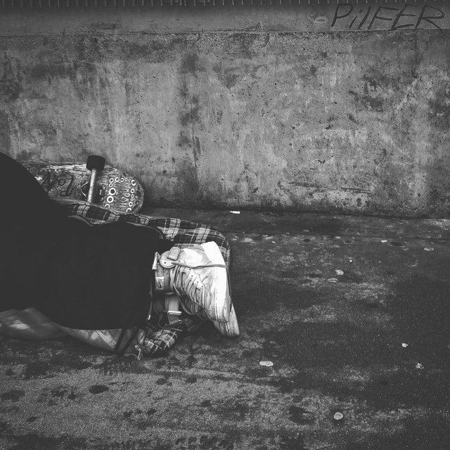 Environmentally Sustainable Travel: A black and white photo of someone's leg's wearing black pants and white sneakers. Behind them is a skateboard. They are sleeping on the street, laying on a plaid sleeping bag.