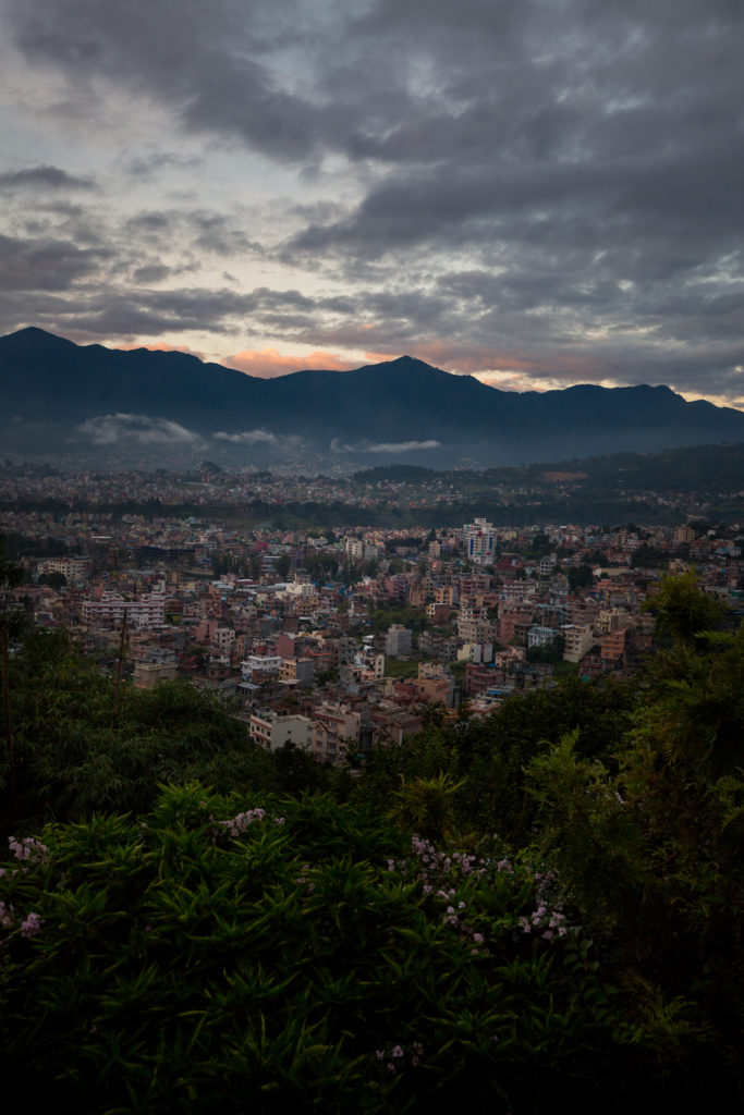 Everest Base Camp Image: A view of the city of Kathmandu, Nepal as seen from the wilderness with mountains seen in the background, peeking from a low-lit sky.