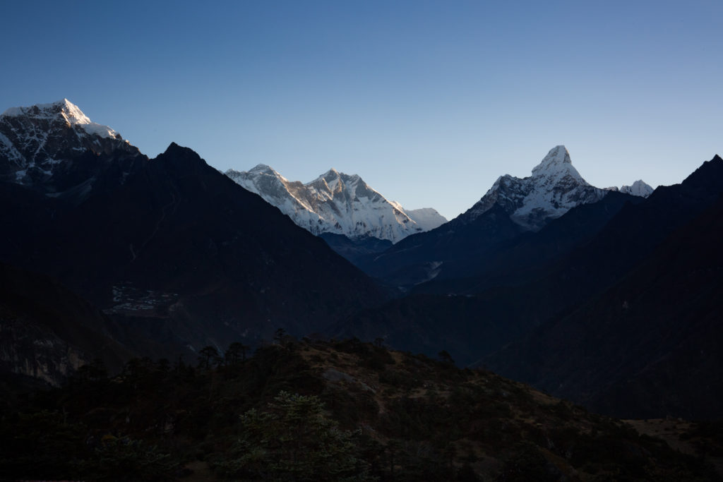 Everest Base Camp Image: A view of snow capped mountains kissed by sunlight rising majestically from amidst smaller mountains in shades of green and brown.