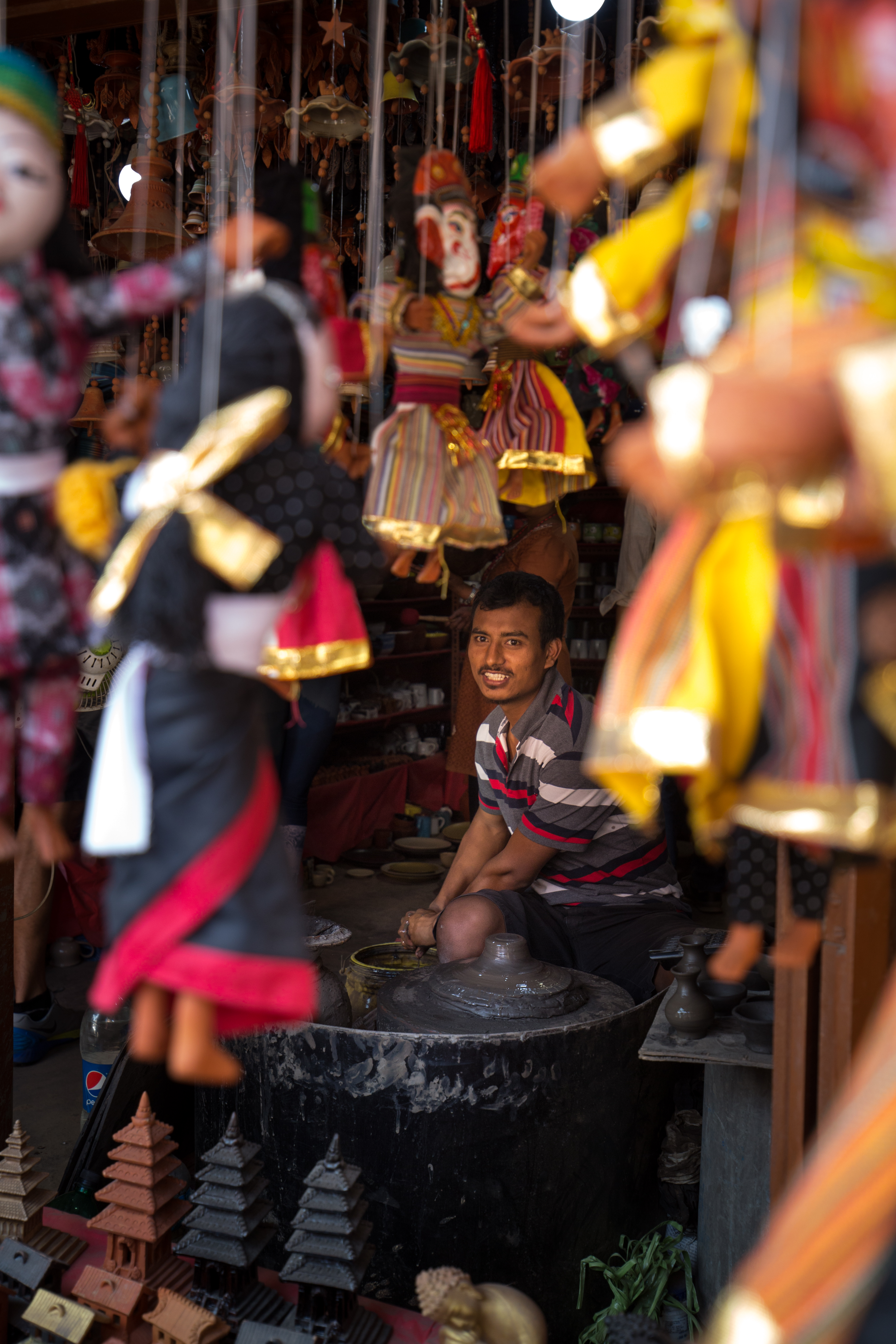 Everest Base Camp Image: An artisan is seen gently smiling from behind a barrage of multicolored puppets suspended from strings, a pottery wheel, and small statues.