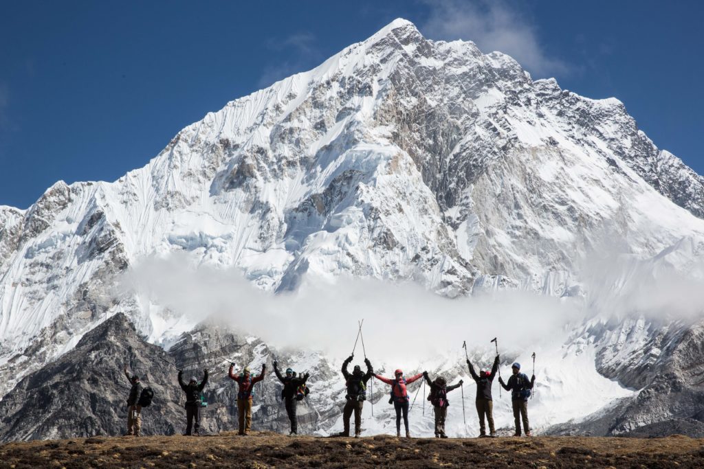 The Everest Base Camp trek team strikes a pose in front of the namesake mountain. The photo is taken at a distance, so it is difficult to make out their individual faces.