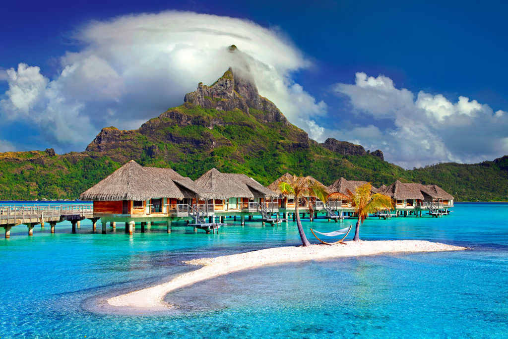 Family Adventure Image: Beach villas built on stilts sit atop crystal clear water. A mountain in shades of brown and green sits in the background, accented by blue sky and white clouds.