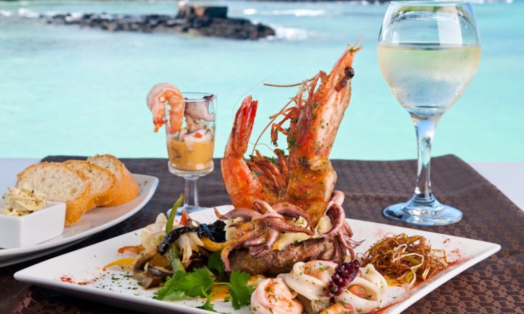 Galápagos Islands Image: A beautiful seafood based meal shows a positioned giant prawn and other delights on a table with an ocean view in the background.