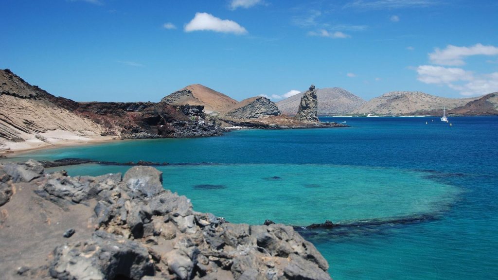 Galápagos Islands Image: A beautiful picture of the Galápagos Islands--the water is turquoise and sapphire, a blue sky is dotted with a few clouds, rocky brown islands and sandy shores can be seen; there is a boat in the distance. 