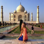 A photograph of author, Hayley Lewis. She wears a blue skirt and orange top. Sitting, smiling at the camera; India's Taj Mahal is in the background.