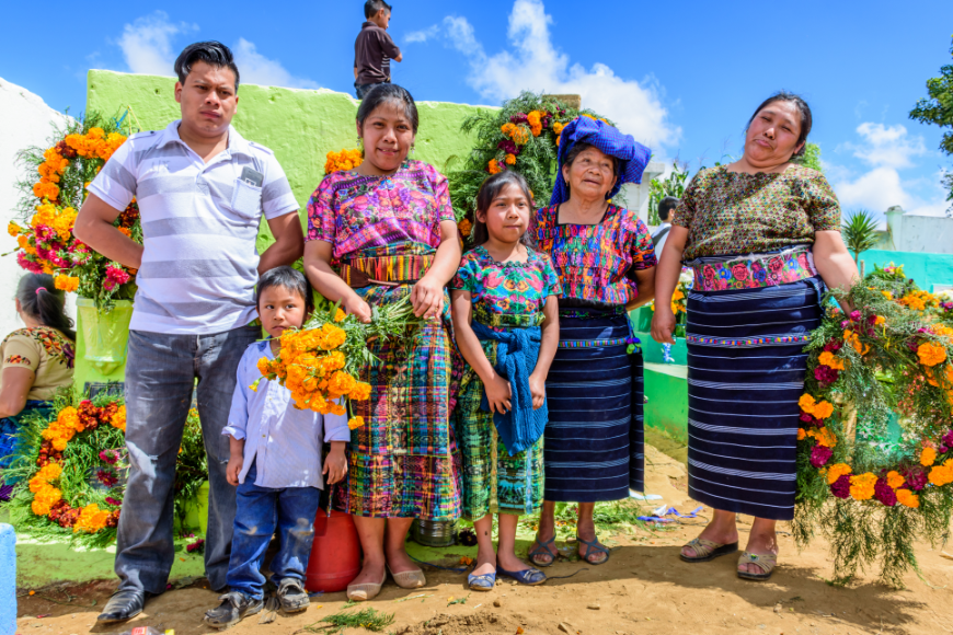 Mayans of Guatemala Image: A family of six (mother, father, son, daughter, aunt, and grandmother) wear bold clothes, hold vibrant flowers, and pose in a colorful Guatemalan cemetry.