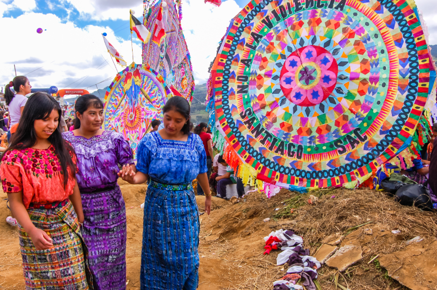 Mayans of Guatemala Image: Three young women in red, purple, and blue pass by colorful, oversized kites.