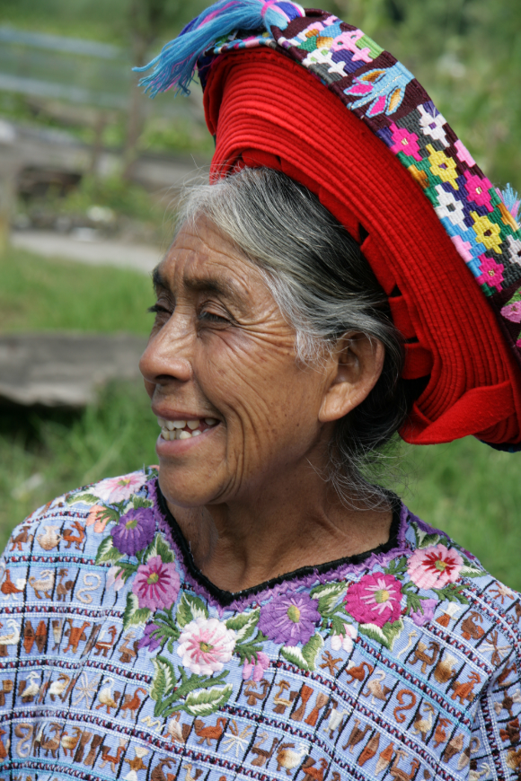 Mayans of Guatemala Image: An elder Guatemalan woman with silver hair smiles wearing an embroidered blouse and red hat.