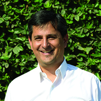 Percy Coll smiles in front of a leafy green background. He is wearing a white button-down shirt. 